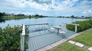 Una terraza de madera con una mesa y sillas da vista a un lago tranquilo, rodeado de follaje y un cielo despejado.