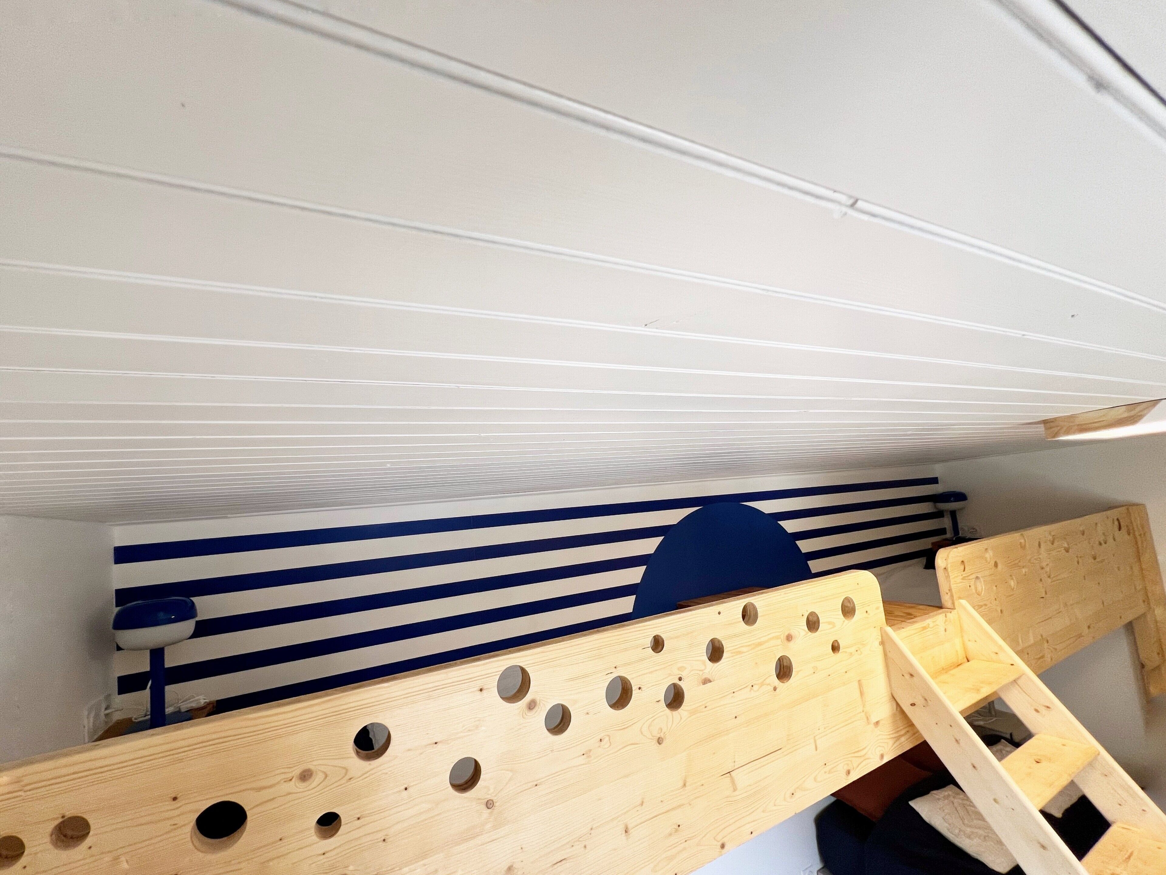 Una stanza con soffitto spiovente, una scala in legno e un muro a righe blu e bianche.