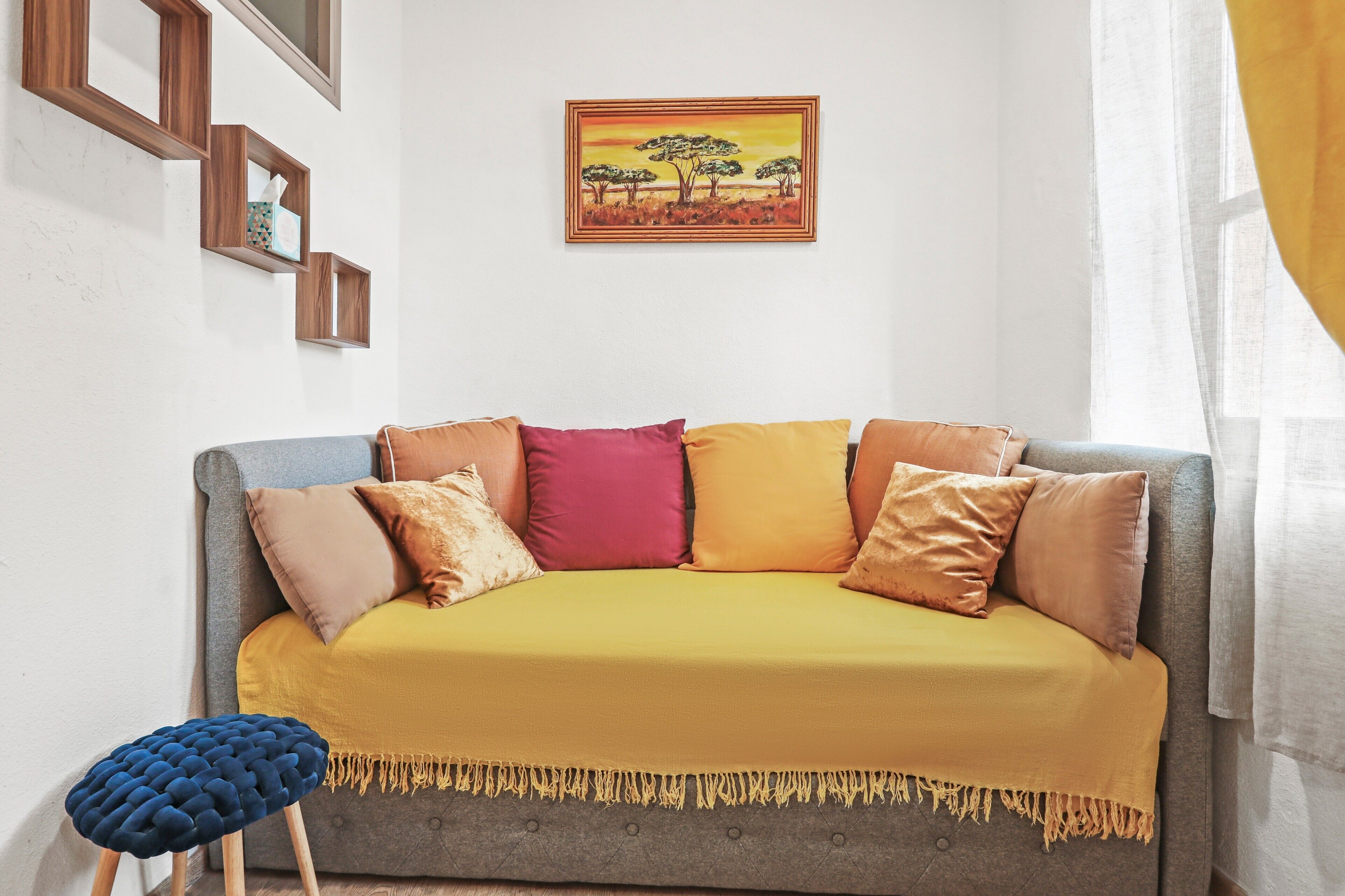 A living room with a sofa, cushions, a small stool, and a framed painting on the wall.
