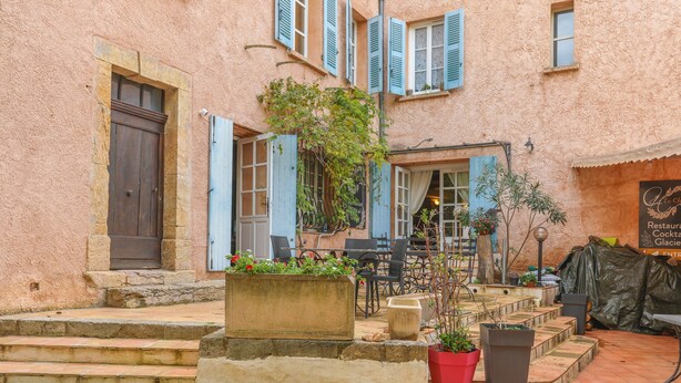 Un bâtiment avec une façade en pierre, des volets bleus, des places assises à l’extérieur et une enseigne indiquant qu’il s’agit d’un restaurant.