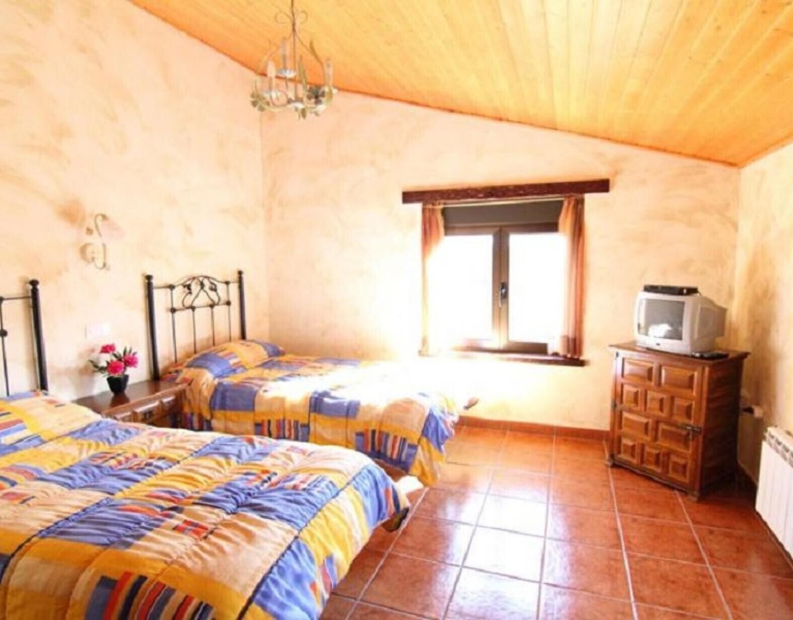 A bedroom with two beds, a wooden ceiling, a window with curtains, a television on a cabinet, and a chandelier.