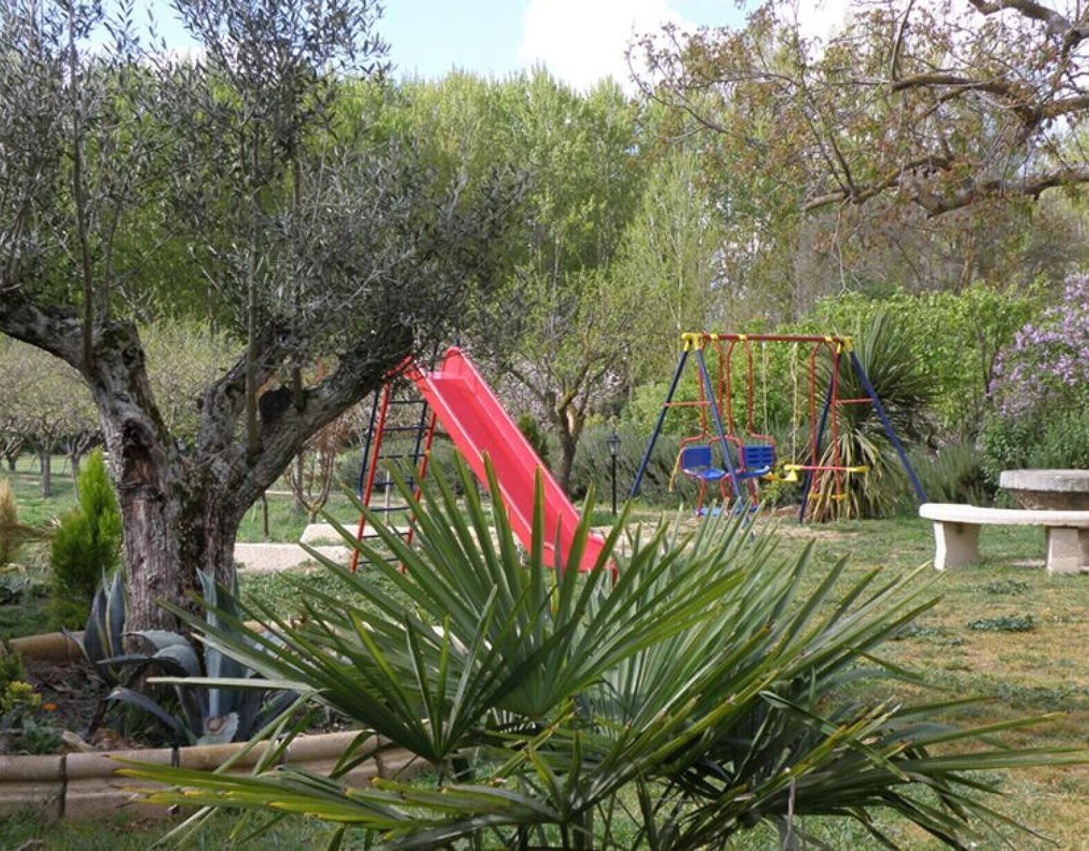 Un parque infantil con un tobogán rojo y columpios, rodeado de árboles y plantas.