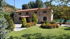 A stone building with a tiled roof, a small porch, and a well-maintained lawn with various shrubs and flowers. - Casa Rural El Manantial near Ciudad Rodrigo with Private Pool and Mountain Views (CIUDAD RODRIGO)