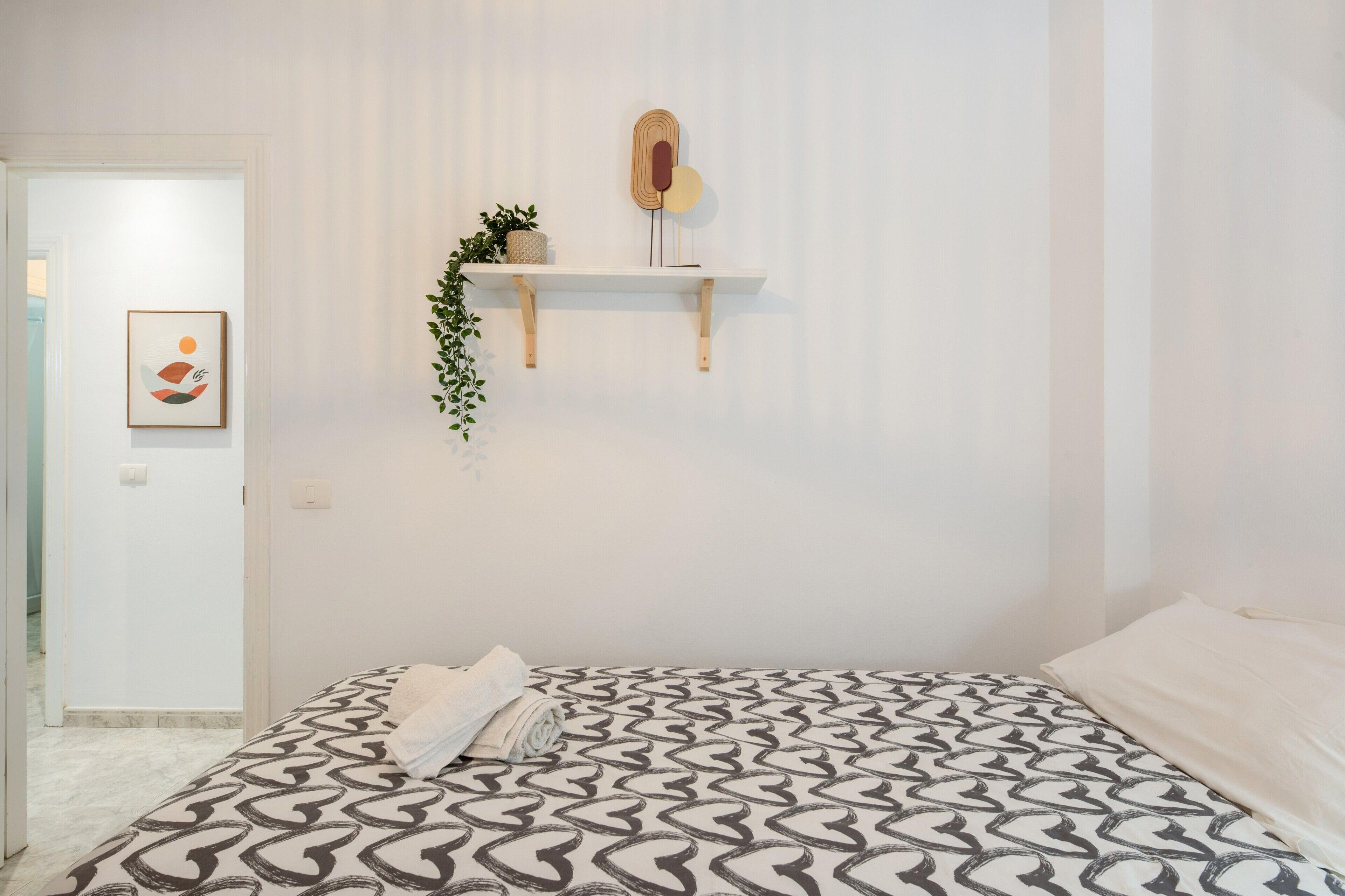 A bedroom with a patterned bedspread, a bedside plant, a wall-mounted shelf with a mirror, and a framed picture.