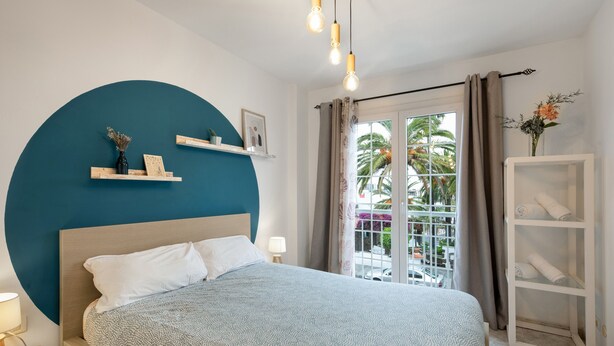 A bedroom with a bed, a large blue wall, a window with curtains, and a view of palm trees. - Apartment 'La Fuente' with Balcony and Wi-Fi (El Paso)