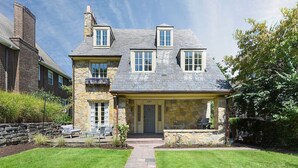 A two-story stone house with a stone chimney, a covered porch, and a stone pathway leading to the entrance. - ★ HostWise Stays ★ Charming Squirrel Hill Home with Garage, Near CMU (Pittsburgh)