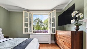 A bedroom with a large bed, a wooden dresser, a TV mounted on the wall, and a bay window with an open shutter. - ★ HostWise Stays ★ Charming Squirrel Hill Home with Garage, Near CMU (Pittsburgh)