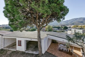 A building with a large tree in front, a patio with chairs, and a view of a town and mountains in the background. - Holiday Home 'Ca La Yaya' with Mountain View, Wi-Fi and Air Conditioning (Llançà)
