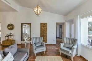 A spacious living room with two grey armchairs, a sofa, a chandelier, a decorative mirror, and a large window with curtains. - Holiday Home 'Ca La Yaya' with Mountain View, Wi-Fi and Air Conditioning (Llançà)