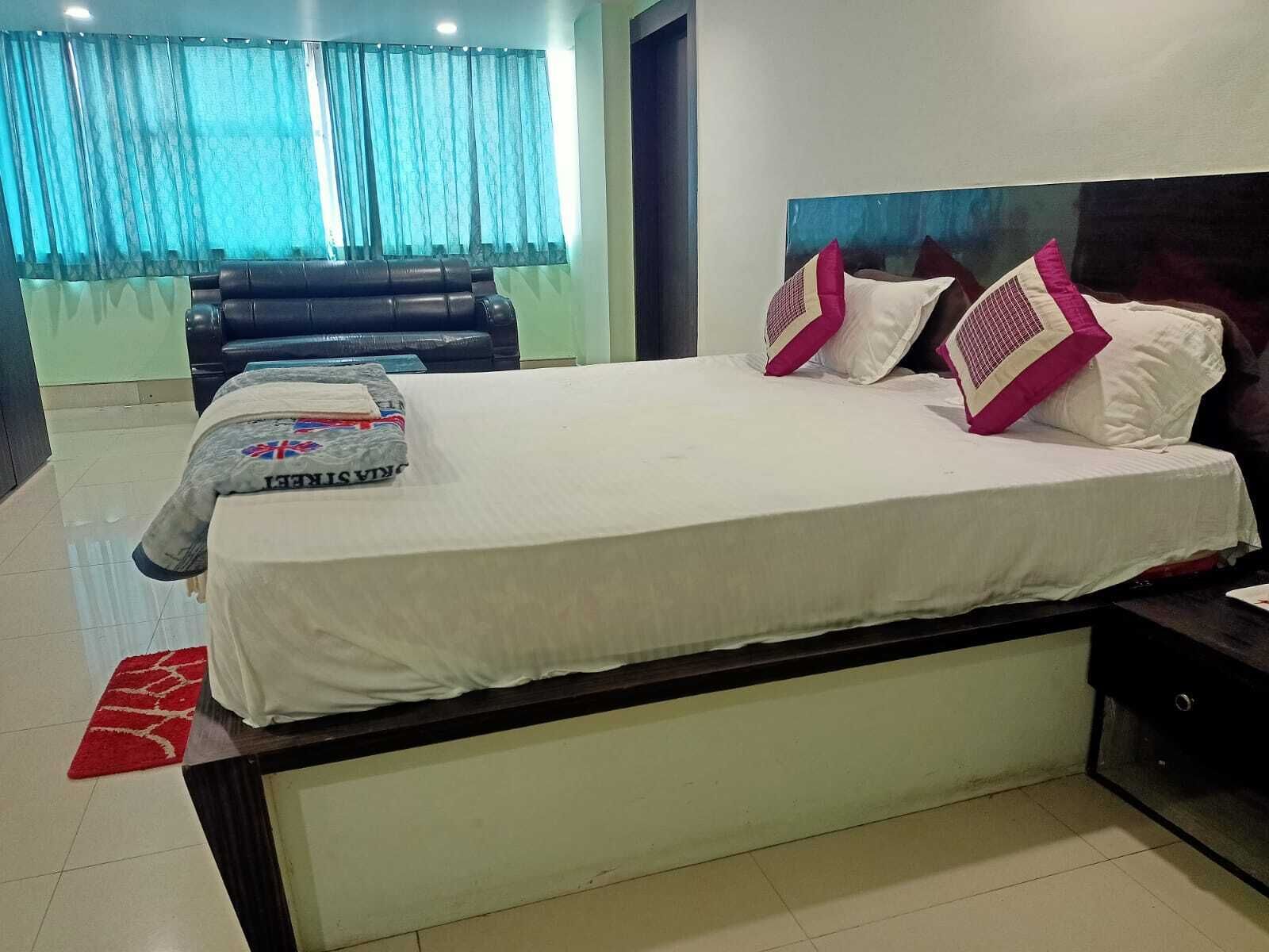 A neatly made bed with white bedding and two decorative pillows, a black leather sofa, and a red rug in a modern hotel room.
