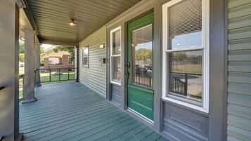 A covered porch with a green door and windows, a metal railing, and a brick building in the background.