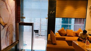 Un hall d’hôtel moderne avec un grand mur en marbre, un ascenseur en verre et un canapé agrémenté de coussins orange.