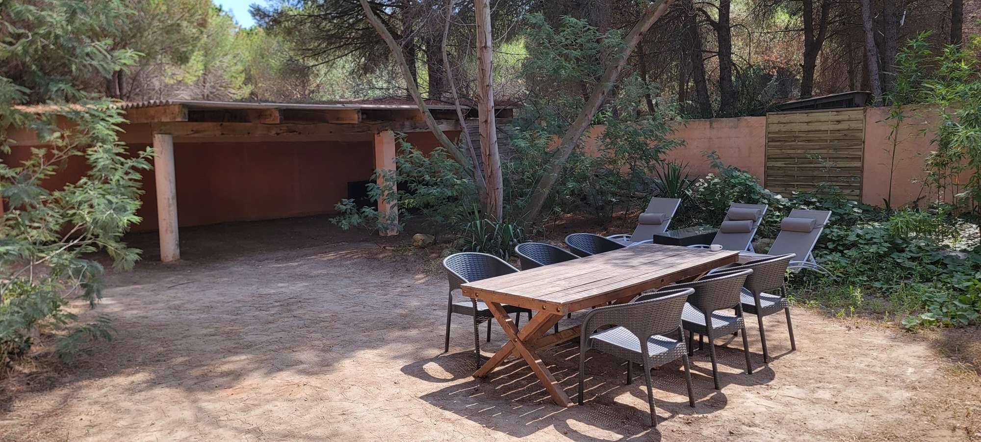 An outdoor patio with a wooden table and wicker chairs, surrounded by a garden and a wooden structure.
