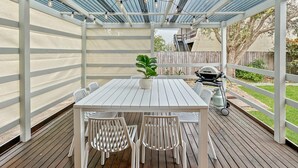 Eine überdachte Terrasse mit einem weißen Tisch und Stühlen, Lichterketten und einem Grill im Hintergrund.