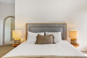 A neatly made bed with a grey headboard, two bedside lamps, and a mirror on the wall. - Beachside 4-Bed Retreat with Study Nook & Views (Coolum Beach)