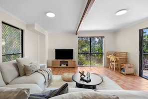 A living room with a sofa, a coffee table, a TV, and a balcony with a view of greenery. - Beachside 4-Bed Retreat with Study Nook & Views (Coolum Beach)