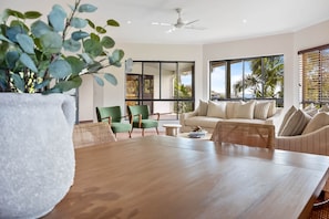 A spacious living room with a wooden table, green chairs, a beige sofa, and large windows offering a view of palm trees.