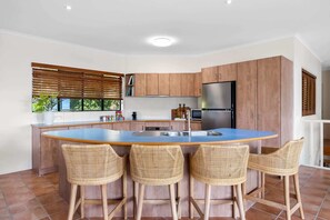 A kitchen with a central island, four wicker chairs, wooden cabinets, and a stainless steel refrigerator. - Beachside 4-Bed Retreat with Study Nook & Views (Coolum Beach)