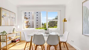 A modern dining area with a glass table, white chairs, and a view of buildings through large windows. - Gorgeous 3-Bed Manly Pad, Short Walk to the Beach (Manly)