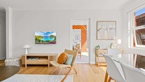 A modern living room with a wooden floor, a white sofa, a wicker chair, and a dining area with a white table and chairs. - Gorgeous 3-Bed Manly Pad, Short Walk to the Beach (Manly)