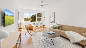 A modern living room with a large window, a ceiling fan, and a dining area with chairs and a table. - Gorgeous 3-Bed Manly Pad, Short Walk to the Beach (Manly)