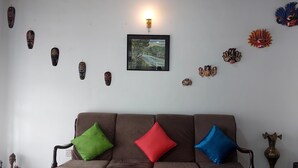 Una sala de estar con un sofá marrón, tres cojines, un cuadro enmarcados de un paisaje, y diversas máscaras decorativas en la pared.