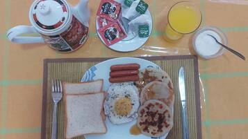 A breakfast plate with sausages, eggs, toast, and pancakes, a mug, tea bags, and a glass of orange juice on a table.