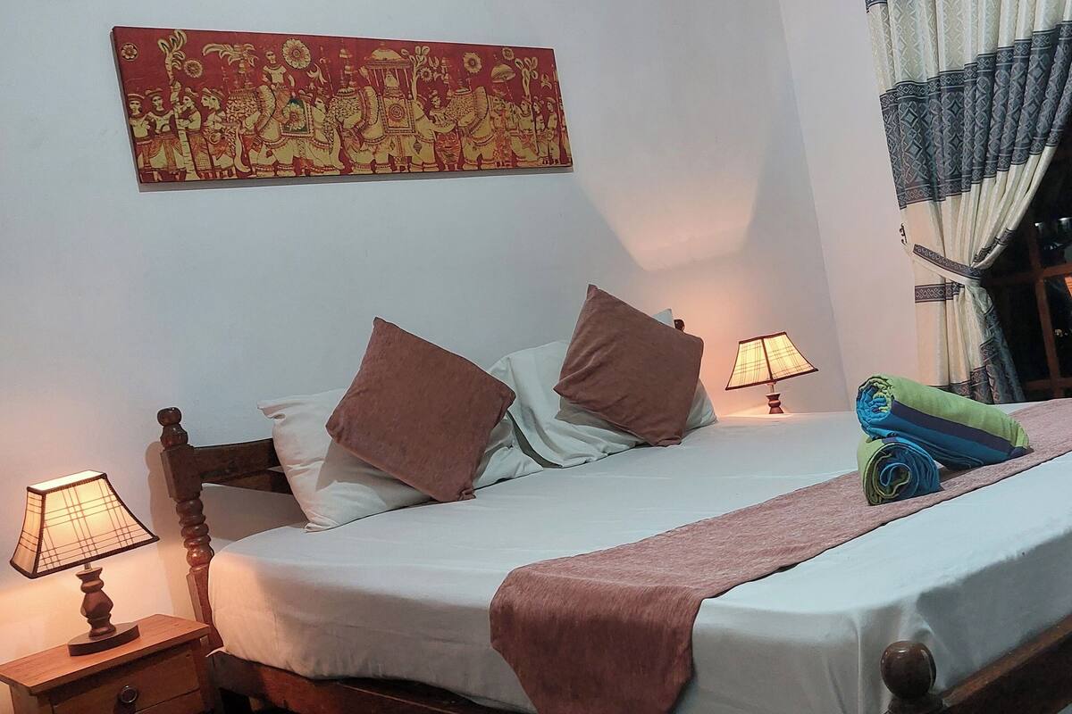 A neatly made bed with a brown blanket, two pillows, and a decorative towel. There are two bedside lamps, a wall hanging, and a window with curtains.