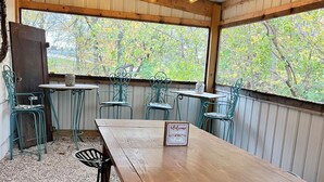 Une table en bois, ornée d’une pancarte « Bienvenue dans la forêt », entourée de chaises en métal vert, sous un toit en tôle ondulée décoré de guirlandes lumineuses.