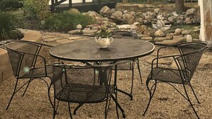 Une terrasse extérieure avec une table ronde et des chaises, des guirlandes lumineuses, une clôture en bois et un petit jardin agrémenté de rochers et de plantes.