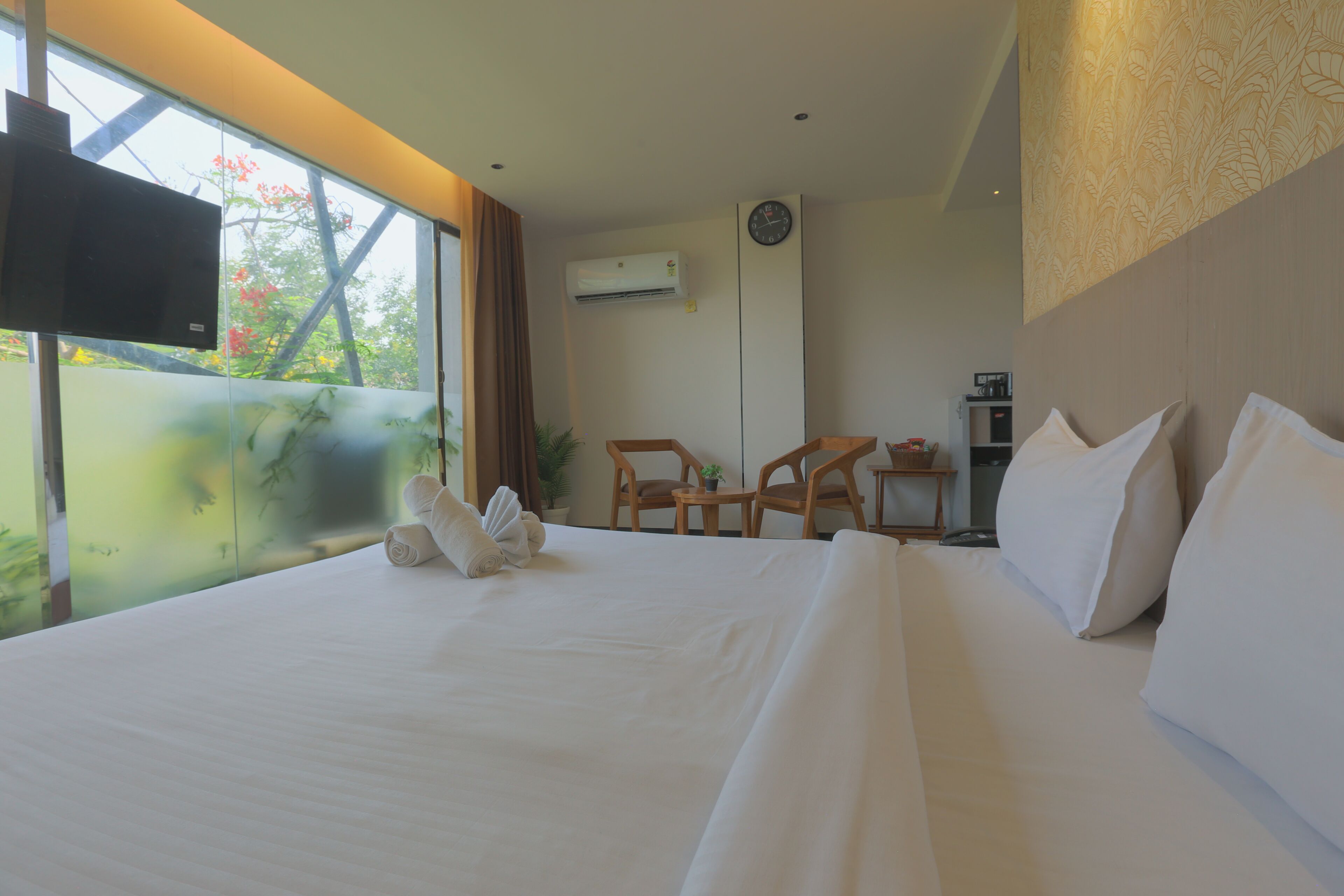 A hotel room with a bed, a TV, a clock, and a view of trees.