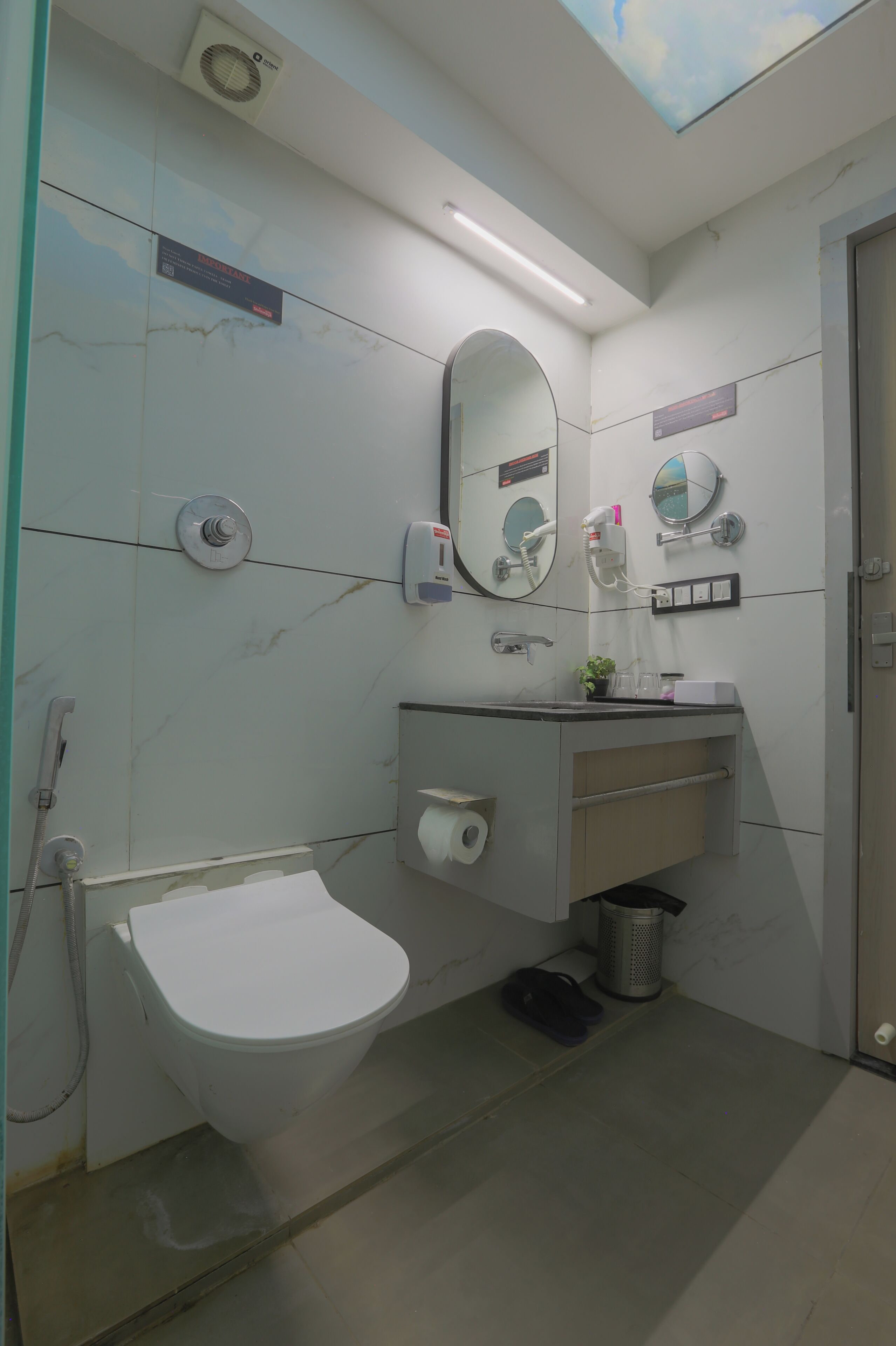 A bathroom with a toilet, a sink, a mirror, a hand dryer, a trash bin, and a ceiling light.