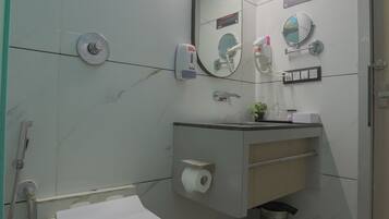 A bathroom with a toilet, a sink, a mirror, a hand dryer, a trash bin, and a ceiling light.