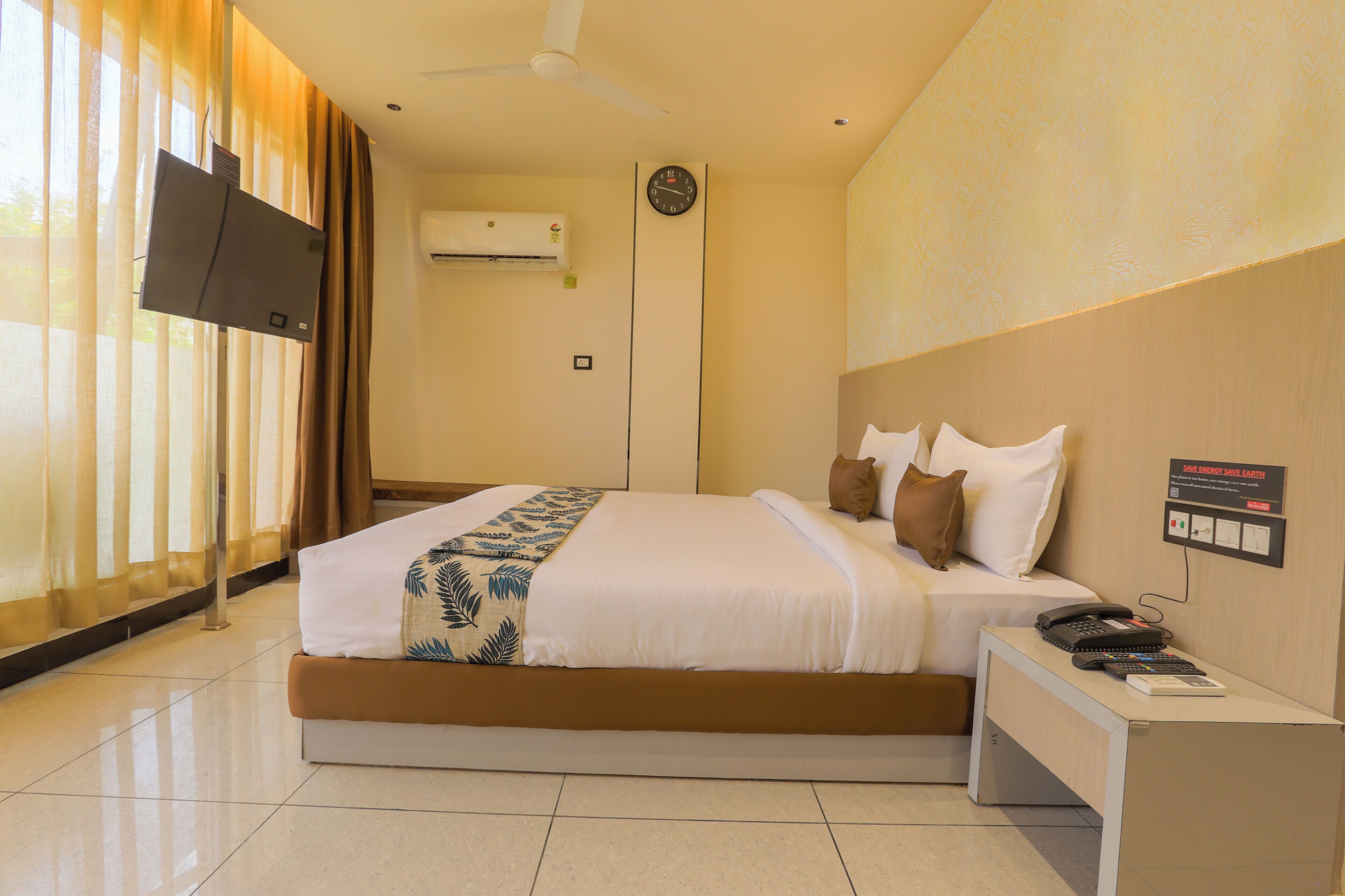 Une chambre d’hôtel équipée d’un lit, d’une table de chevet avec un téléphone, d’une horloge murale, d’un climatiseur et d’un téléviseur fixé au mur.