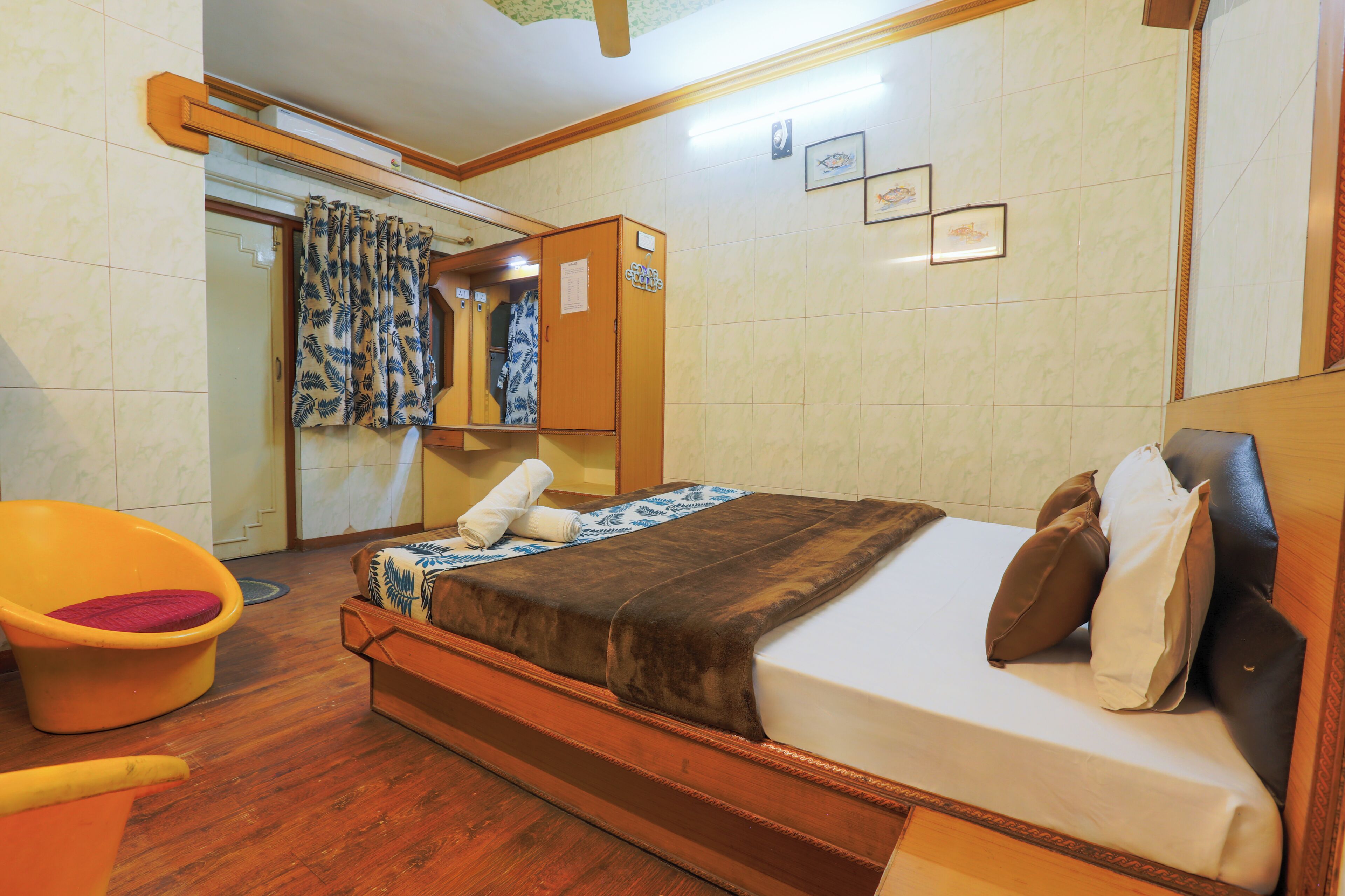 Une chambre d’hôtel avec un lit, une tête de lit en bois, une armoire en bois, une salle de bain avec un rideau de douche et une chaise jaune.