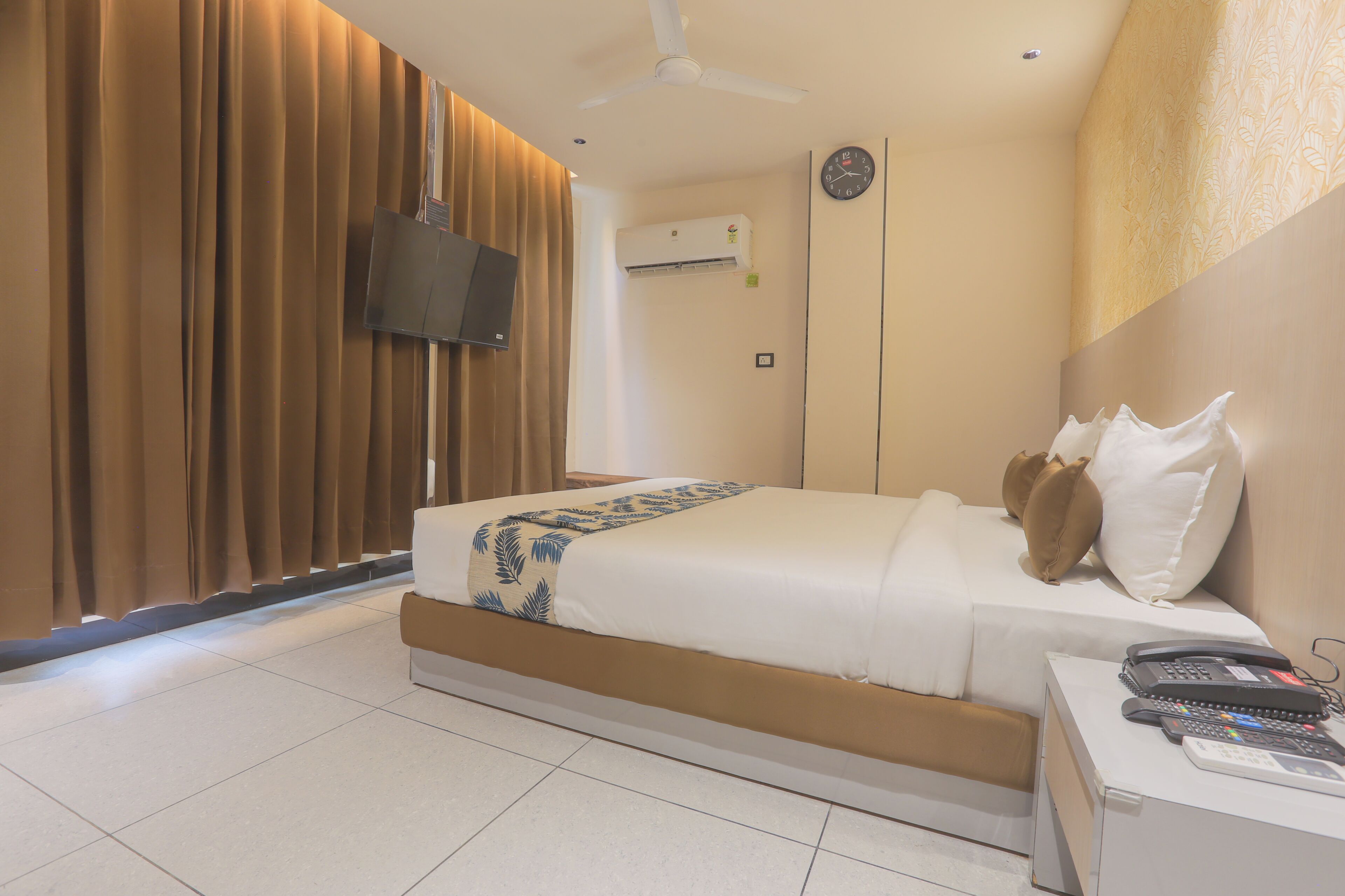 A hotel room with a bed, a bedside table with a phone and remote controls, a wall-mounted TV, and an air conditioning unit.