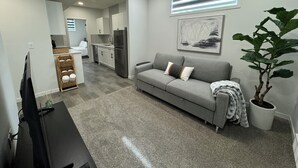 Ein modernes Wohnzimmer mit einem grauen Sofa, einem Flachbildfernseher, einer Zimmerpflanze und einer Küche im Hintergrund.