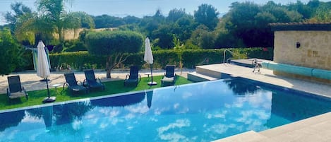 A clear blue swimming pool with lounge chairs and umbrellas, surrounded by greenery and a stone building.