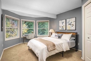 A bedroom with a large bed, two windows with a view of trees, a desk with a chair, and two abstract paintings on the wall.