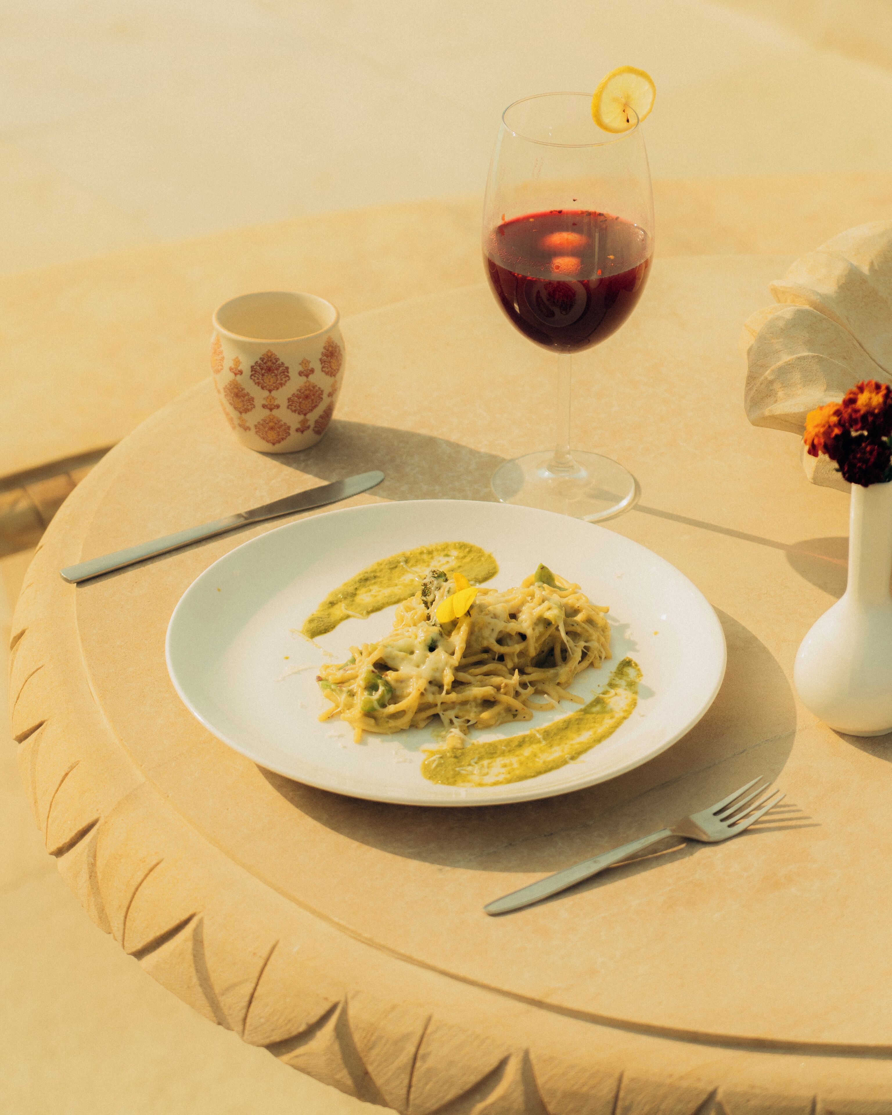 A plate of pasta with a lemon garnish, a glass of red wine, a floral cup, and a vase with flowers on a round table.
