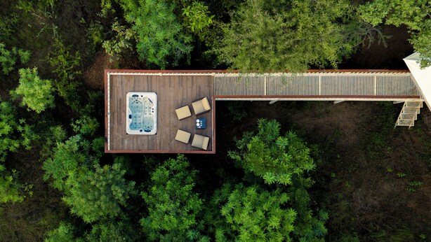 An aerial view of a wooden deck with a hot tub and seating area surrounded by dense greenery.