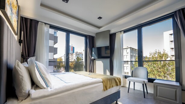A modern hotel room with a bed, a chair, a TV, and large windows offering city views. - Alion Hotel (Izmir)
