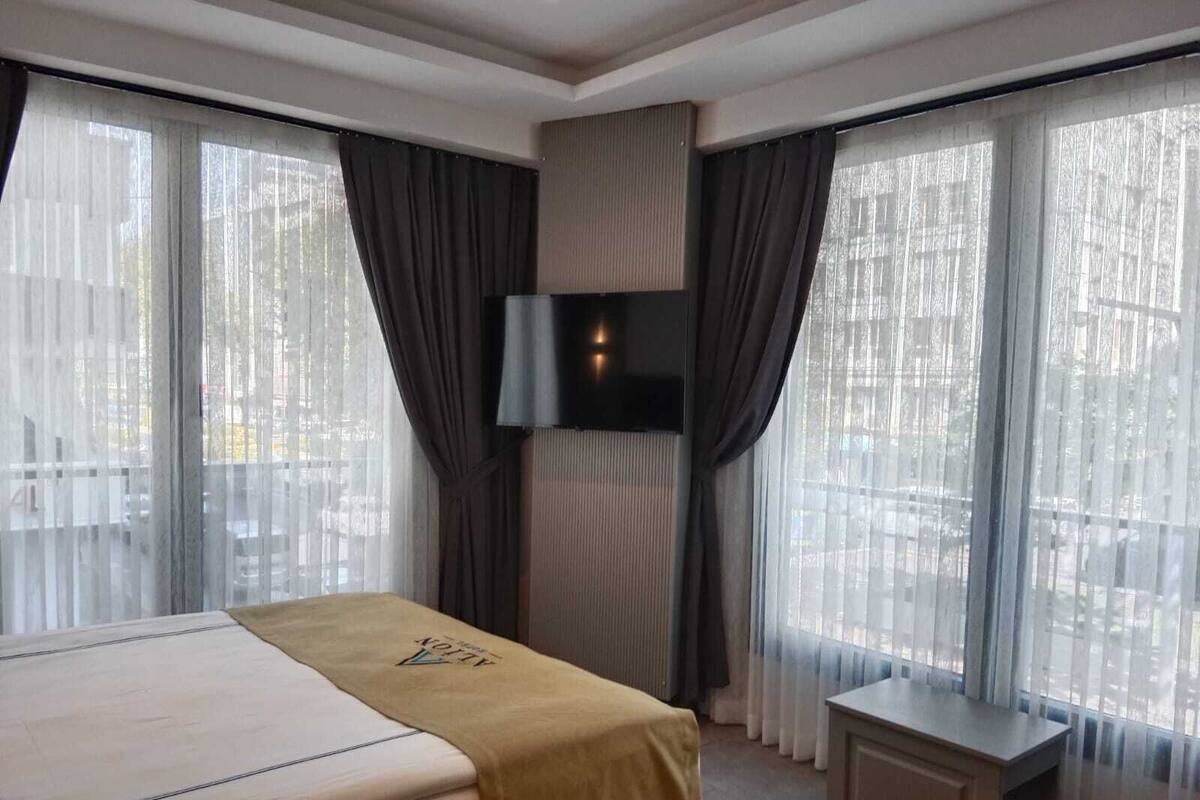 Ein Hotelzimmer mit einem Bett, einem an der Wand befestigten Fernseher und großen Fenstern mit Vorhängen.