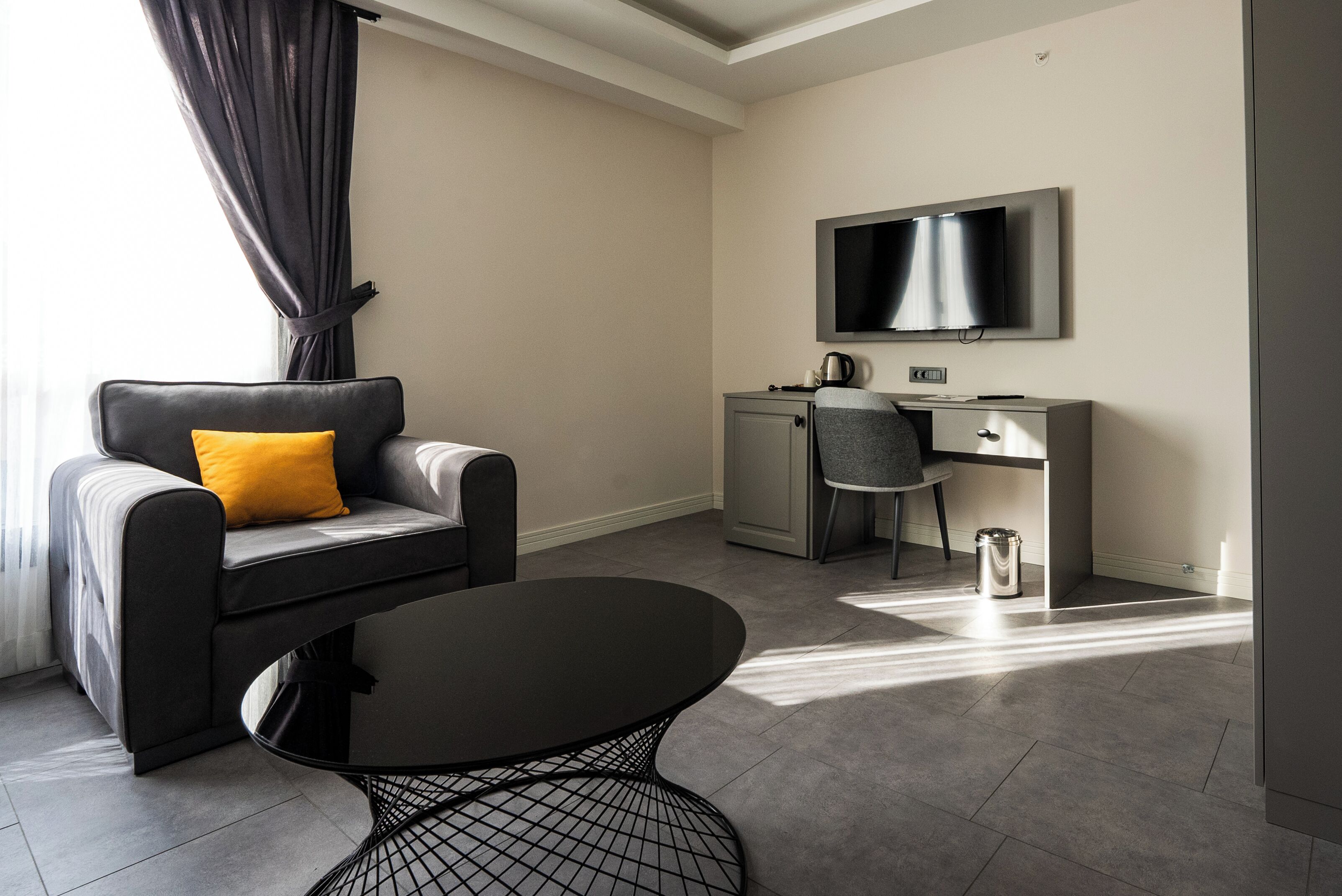 A modern hotel room with a sofa, a round table, a TV mounted on the wall, and a small kitchenette area.