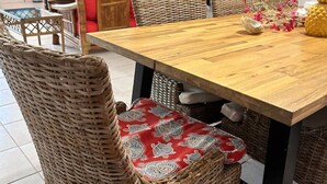A dining area with a wooden table and wicker chairs, a black bookshelf, and a red sofa in the background. - Travelers Palm Beachy Paradise (Edgewater)
