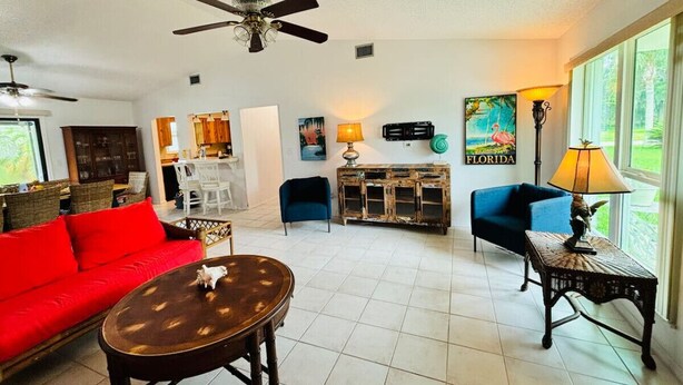 A spacious living room with a red sofa, a coffee table, and a blue armchair. There is a dining area in the background with a white table and chairs. - Travelers Palm Beachy Paradise (Edgewater)