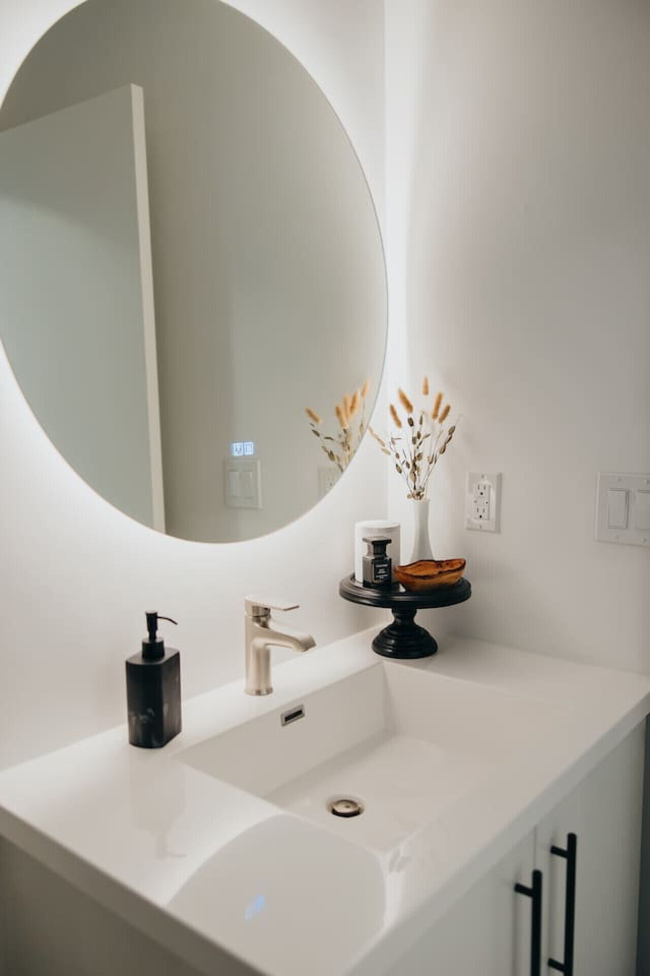Un bany modern amb un mirall rodó gran, un lavabo blanc, un dosificador de sabó negre i un vas petit amb flors seques.