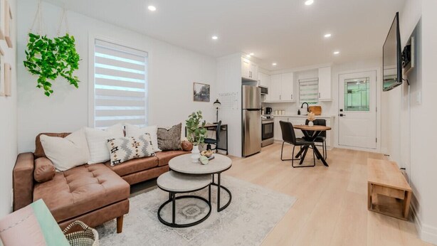 A modern apartment with a living room, dining area, and kitchen. It features a brown leather sofa, a round coffee table, and a dining table with chairs. The kitchen has stainless steel appliances and white cabinets. - Stylish 1BD Steps from Downtown (Kitchener)