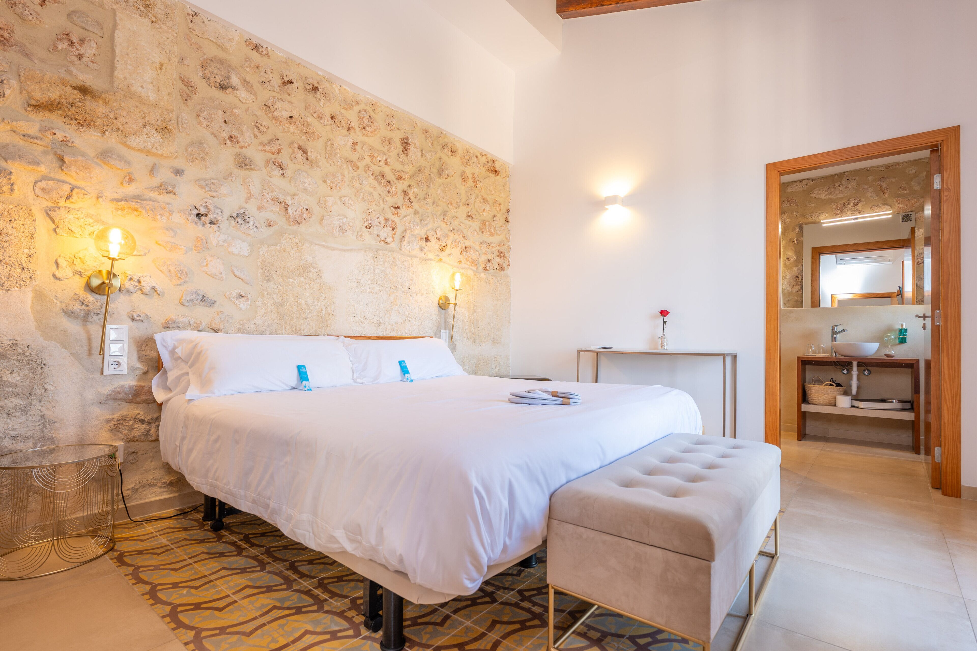 A bedroom with a stone wall, a bed with white bedding, a bedside table, a bench, and a bathroom with a sink and mirror.