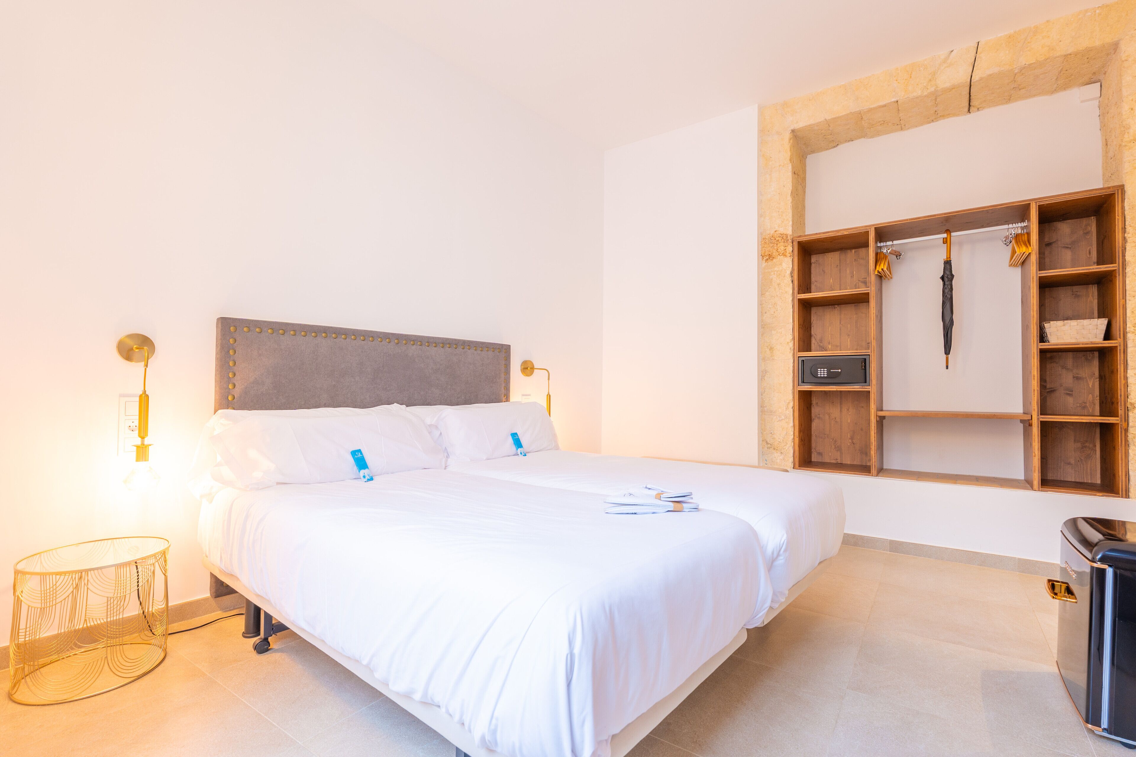 A neatly made bed with white linens, a bedside lamp, and a small round table. There is a wooden shelf unit with a hanging umbrella and a trash bin.